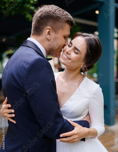 Couple of loving man and woman at wedding ceremony hugging and kissing outdoors. Attractive brides enjoying the moment and smiling. Happiness. Tenderness. Wedding dress. Wedding suit.