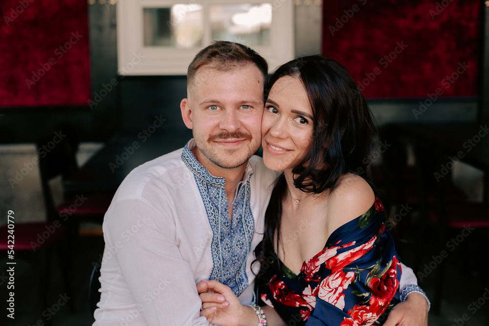 Fototapeta premium Portrait of caucasian couple man and woman embracing man looking at the camera. Happy man in shirt with blue embroidery embracing his attractive girlfriend with long hair, dressed in elegant dress, 