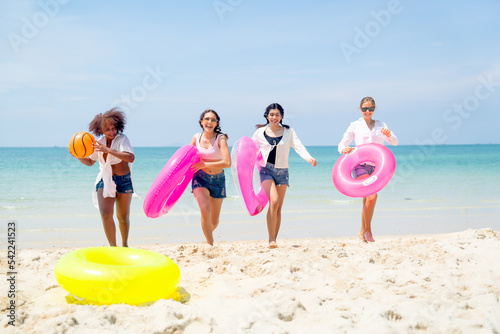 Group of teenage girls hold multicolor swim ring and ball walk back from the sea to beach with blue clear sky as background and they look happy to enjoy during holiday together.