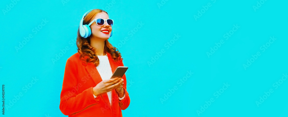Portrait of happy smiling modern young woman in wireless headphones listening to music with smartphone wearing red jacket on blue background