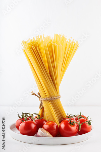 Bundle of raw spaghetti tied with rope stands on table. Dry pasta with cherry tomatoes and garlic on a white plate. Close-up