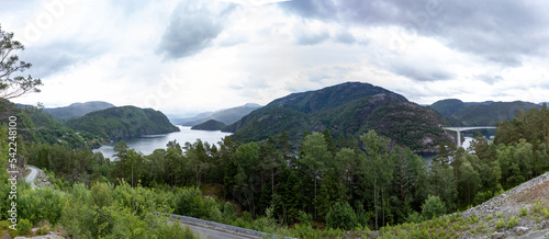 Panoramic view of Norwegian fjord seawater inlet between green mountains with tall vegetation and infrastructure of roads and bridges. 