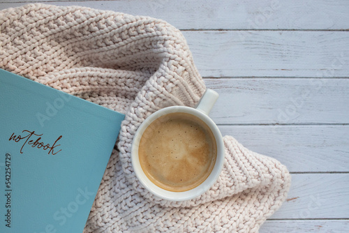 Cup of coffee on wooden table. Hot drink cappuccino in mug, blue notebook and sweater on white background. Cozy winter concept. Top view, flat lay, copy space