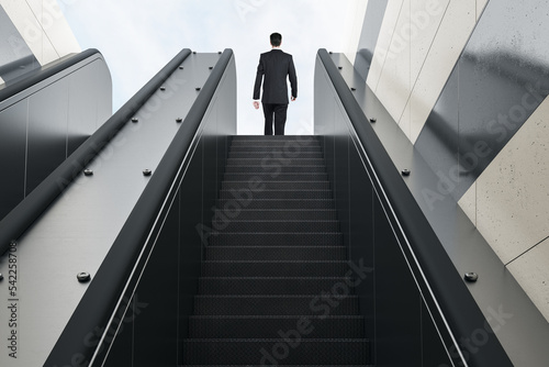 Business success concept with man in black suit back view on the top of escalator moving up on blue sky background