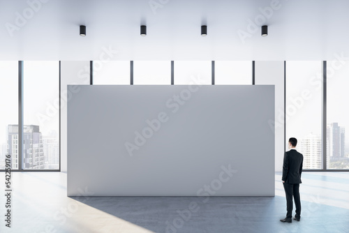 Businessman perspective view looking at big blank white partition with place for advertisement or poster in sunlit spacious empty hall with concrete floor and city view from panoramic windows, mock up