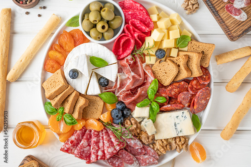 Charcuterie board with a variety of cheeses, salami, chorizzo, prosciutto, honey, grapes, nuts, olives, bread, blueberries and fresh herbs on a white wooden background. A festive snack.