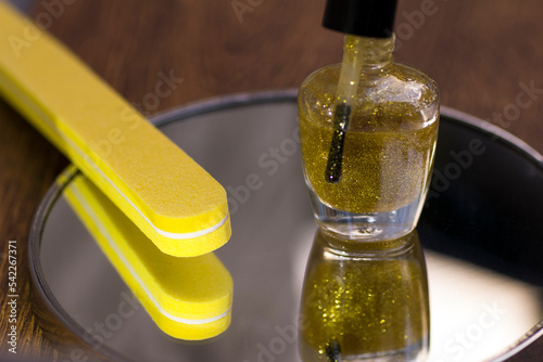 A bottle of translucent nail polish with gold glitter and a yellow nail file on a mirror surface
