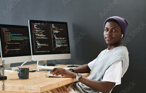 Young black man in casualwear keeping hands on computer keyboard while sitting in front of monitors and looking at camera photo