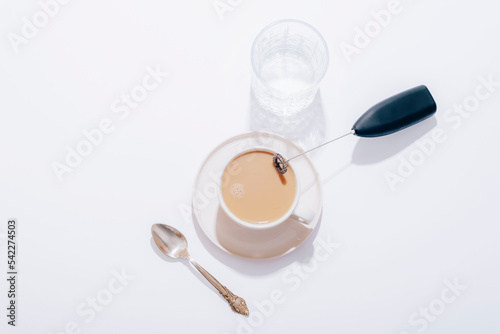 Coffee cup, glass of water, spoon and black milk frother on white table. Top view, flat lay photo