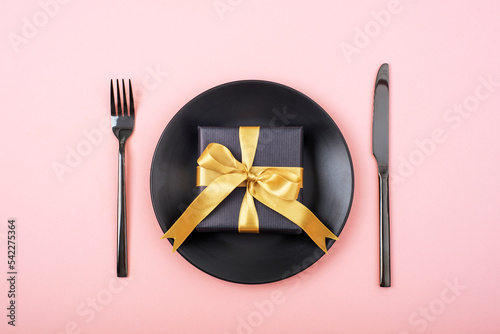Stylish table setting, gift box, black plate, cutlery on pink background. Christmas concept. Top view, flat lay