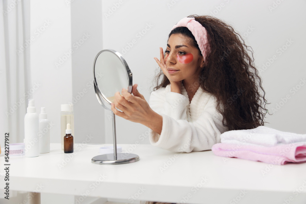 Happy tanned pretty curly Latin lady with pink patch for eye looks at mirror doing beauty procedure in white home interior. Copy space. Moisturizing beauty grooming, skincare and pampering concept Happy tanned pretty curly Latin lady with pink patch for eye looks at mirror doing beauty procedure in white home interior. Copy space. Moisturizing beauty grooming, skincare and pampering concept