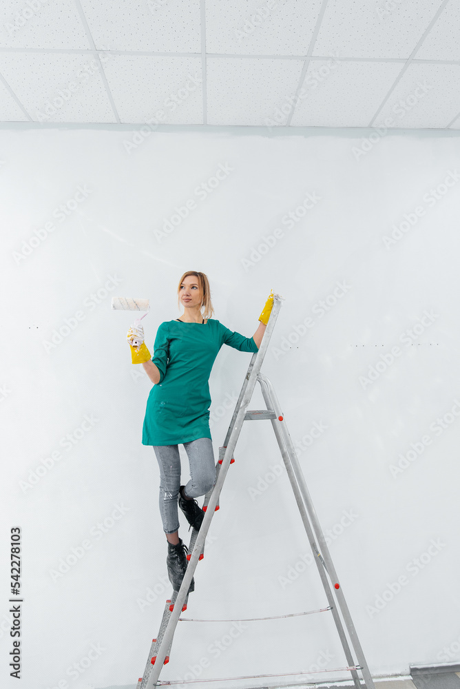A young girl is engaged in repairs on the stairs and paints a white wall with a roller in a new apartment. Renovation of the interior and a new apartment. Housewarming and a desirable mortgage.