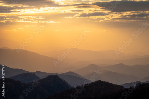 A glowing mountain sunset in the Smoky Mountains taken from the Blue Ridge Parkway. There are clouds in the sky and the hazy atmosphere is glowing orange. There are layers of mountains. 