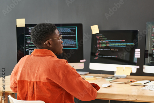 Rear view of young black man concentrating on decoding data on computer screen while sitting by workplace in front of monitors