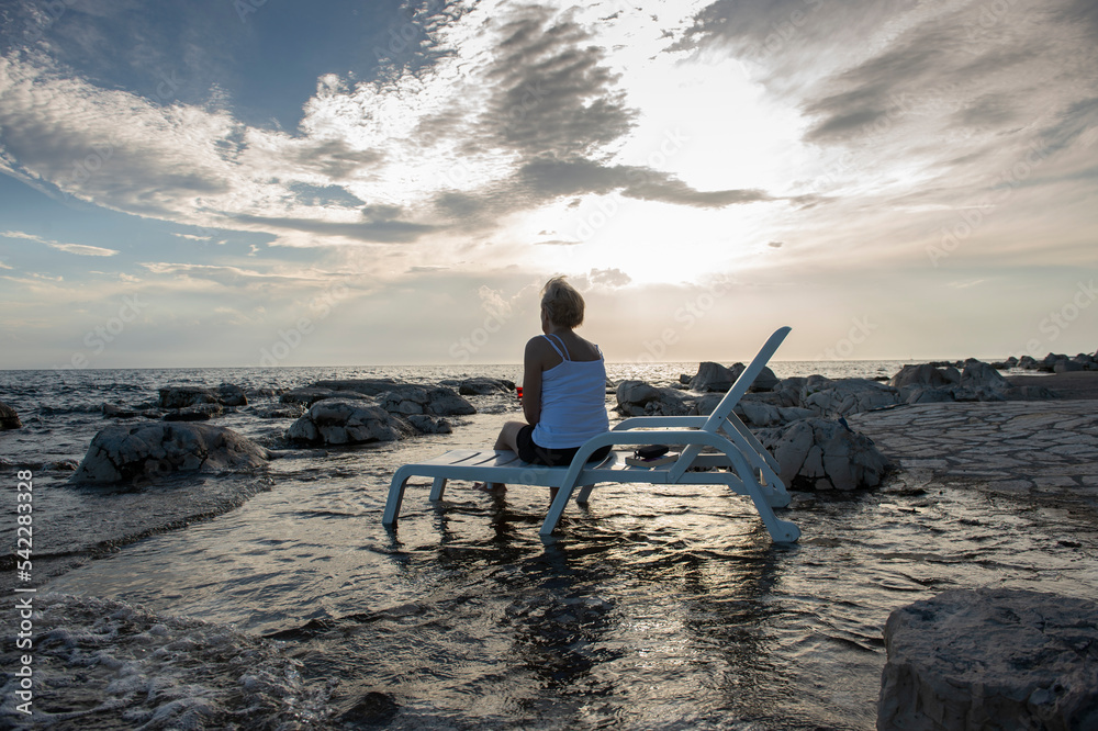 A woman sits on a deck chair on the seashore drinking a drink and enjoying the sunset