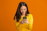 Young Asian woman holding a mobile phone, typing a message, online chatting. Smiling female playing games using smartphone standing on a orange background