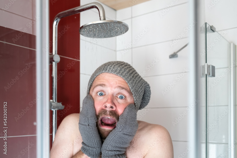 Freezing man in a hat and mittens in the shower under cold water. Concept of energy crisis. - obrazy, fototapety, plakaty 