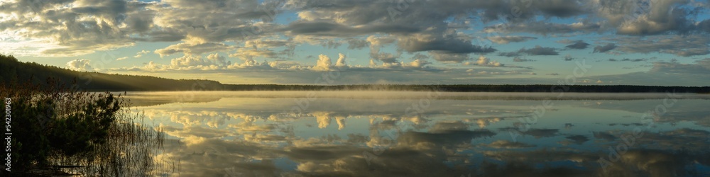 Obraz premium serene morning landscape. the cloudy sky is reflected on the surface of the water with a light atmospheric haze and the warm light of the dawn of the rising sun. widescreen panorama