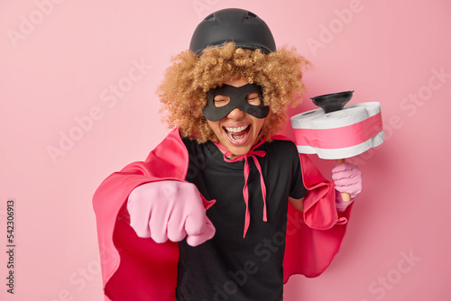 Responsible curly superheroine clenches fist wants to fly and help you to do domestic chores holds plunger with toilet paper around wears protective hemet and cloak isolated over pink background photo