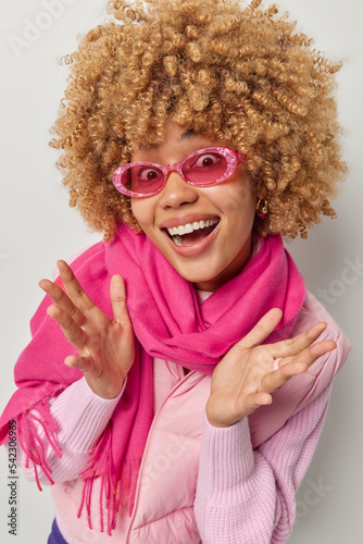 Beautiful cheerful European woman with curly hair spreads palm and looks happily at camera wears pink sunglasses scarf around neck going to have walk in park glad to meet friend poses indoor