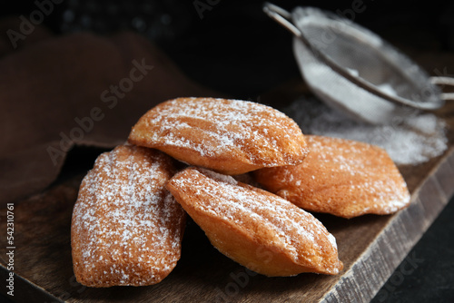 Delicious madeleine cakes with powdered sugar on wooden board, closeup