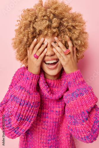 Overjoyed curly haired young European woman covers eyes with hand laughs happily smiles broadly hides her genuine emotions wears knitted jumper isolated over pink background. Happiness concept photo