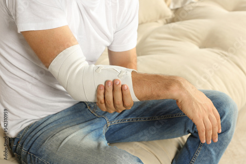 Fototapeta Naklejka Na Ścianę i Meble -  Man with arm wrapped in medical bandage on sofa, closeup