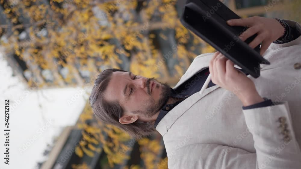Bearded businessman standing at the street and tapping with stylus on gadget computer. Adult boss director using tablet device and browsing online outdoors Bearded businessman standing at the street and tapping with stylus on gadget computer. Adult boss director using tablet device and browsing online outdoors