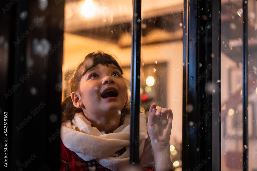 Adorable child look through the window and admiring first snow flakes.