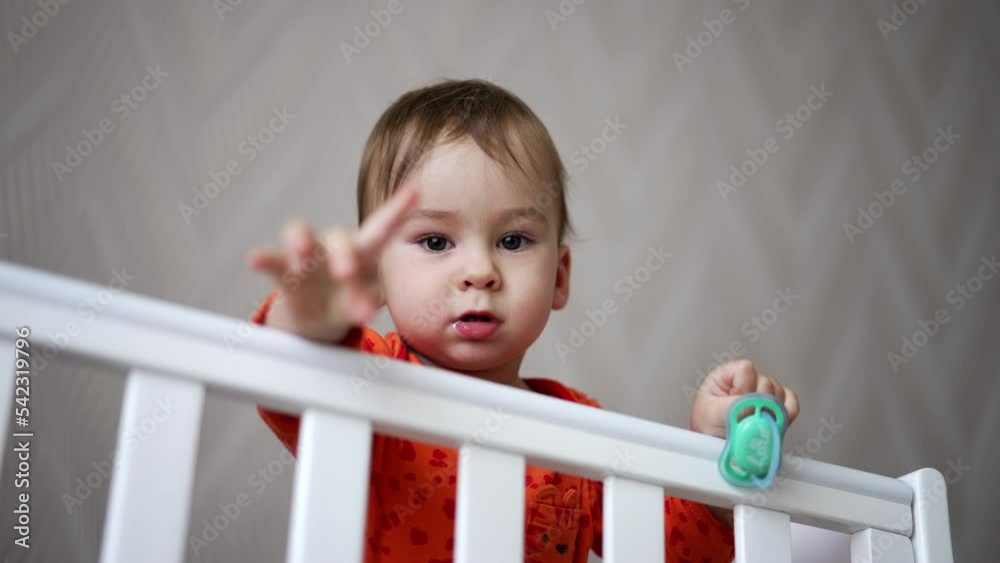 Cute one-year-old child standing in the crib and holding a pacifier in his hand. Baby looks straight into camera showing gestures with his hand. Cute one-year-old child standing in the crib and holding a pacifier in his hand. Baby looks straight into camera showing gestures with his hand.