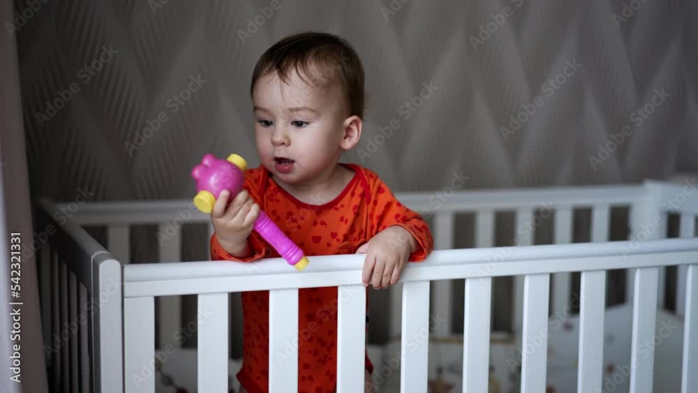 Female hand gives a toy to a baby standing in the crib. Sweet little baby takes a toy and shoves it to his mouth looking up. Close up. Female hand gives a toy to a baby standing in the crib. Sweet little baby takes a toy and shoves it to his mouth looking up. Close up.