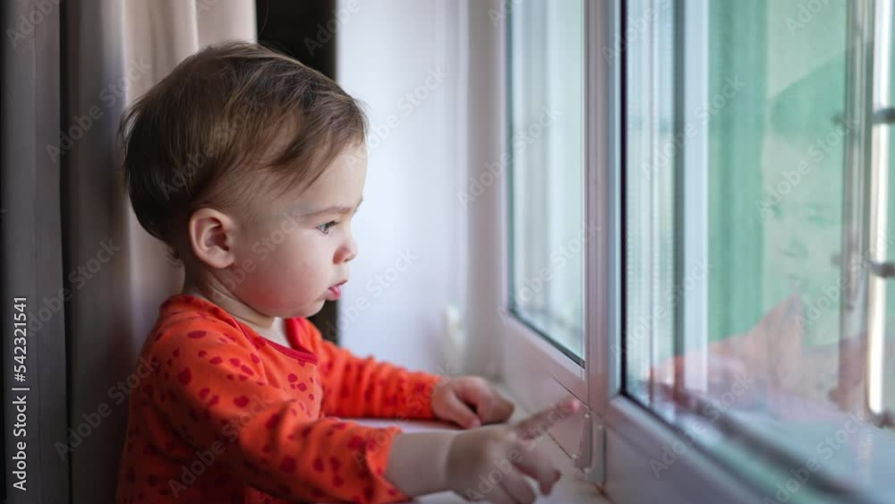 Curious toddler in orange shirt touching the window frame detail with his finger. Baby boy looks into window touching his head. Curious toddler in orange shirt touching the window frame detail with his finger. Baby boy looks into window touching his head.