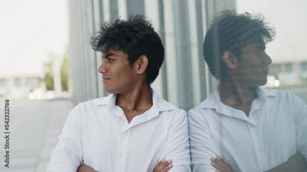 Indian young guy crossing his arms, leaning against a glass wall on the street Indian young guy crossing his arms, leaning against a glass wall on the street