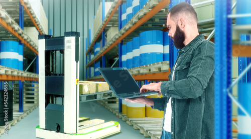 Warehouse technologies. Man with laptop in warehouse building. Robotic forklift concept. Guy is developing robotic forklift. Man and warehouse racks. Robotic forklift technology. Art blurred