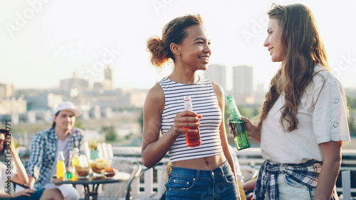 Canvas Print Pretty African American girl is talking to her cheerful Caucasian friend and drinking cocktail holding bottle during open-air party on rooftop