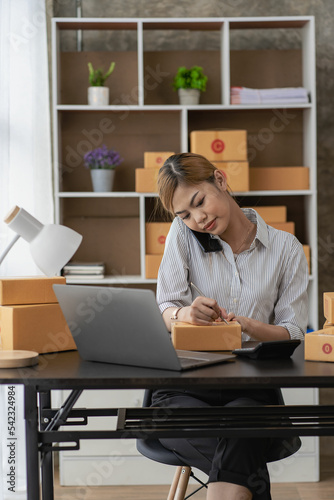  Smiling Asian woman looking at camera and using laptop to check package delivery information before sending it to customer. Small sme business entrepreneur. Online selling concept.