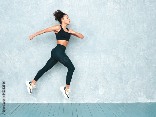 Portrait of fitness smiling black woman in sports clothing with afro curls hairstyle.Wearing sportswear.Sexy young beautiful model doing lunges before training. Female jumping in studio near grey wall