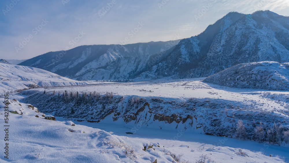 A frozen river winds in a valley between steep banks. Fluffy hoarfrost on dry grass and on the ground. A picturesque mountain range against the blue sky. Altai