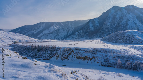 A frozen river winds in a valley between steep banks. Fluffy hoarfrost on dry grass and on the ground. A picturesque mountain range against the blue sky. Altai