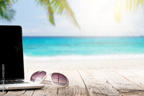 Laptop and sunglasses on wooden table in front of sunny sea