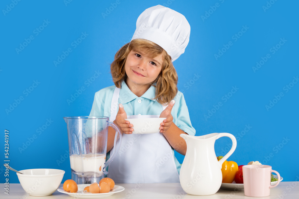 Child chef cook prepares food in isolated blue studio background. Kids cooking. Teen boy with apron and chef hat preparing a healthy vegetables meal in the kitchen. Child chef cook prepares food in isolated blue studio background. Kids cooking. Teen boy with apron and chef hat preparing a healthy vegetables meal in the kitchen.