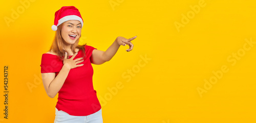 Cheerful young Asian woman wearing christmas hat pointing finger at camera over yellow background