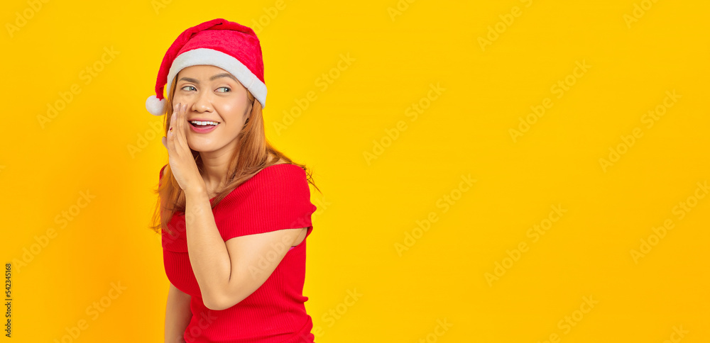 Cheerful young Asian woman wearing Christmas hat whispering something on yellow background Cheerful young Asian woman wearing Christmas hat whispering something on yellow background