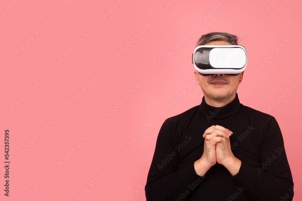 Portrait of Asian man wearing a black turtleneck using virtual reality headset praying on pink background. Free from copy space. VR, future, gadgets, technology, studying, video game concept Portrait of Asian man wearing a black turtleneck using virtual reality headset praying on pink background. Free from copy space. VR, future, gadgets, technology, studying, video game concept