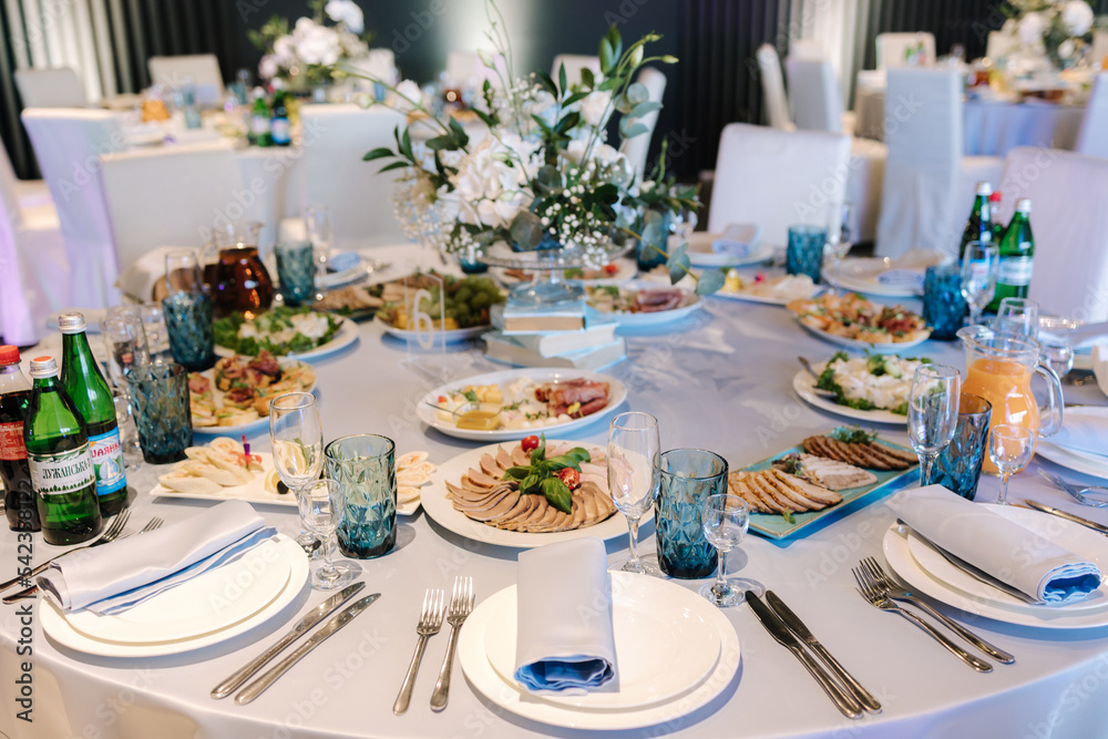 Big round table decorated by white and blue flowers in restaurant. Different dishes on table. Cristening of little boy. Luxury decor