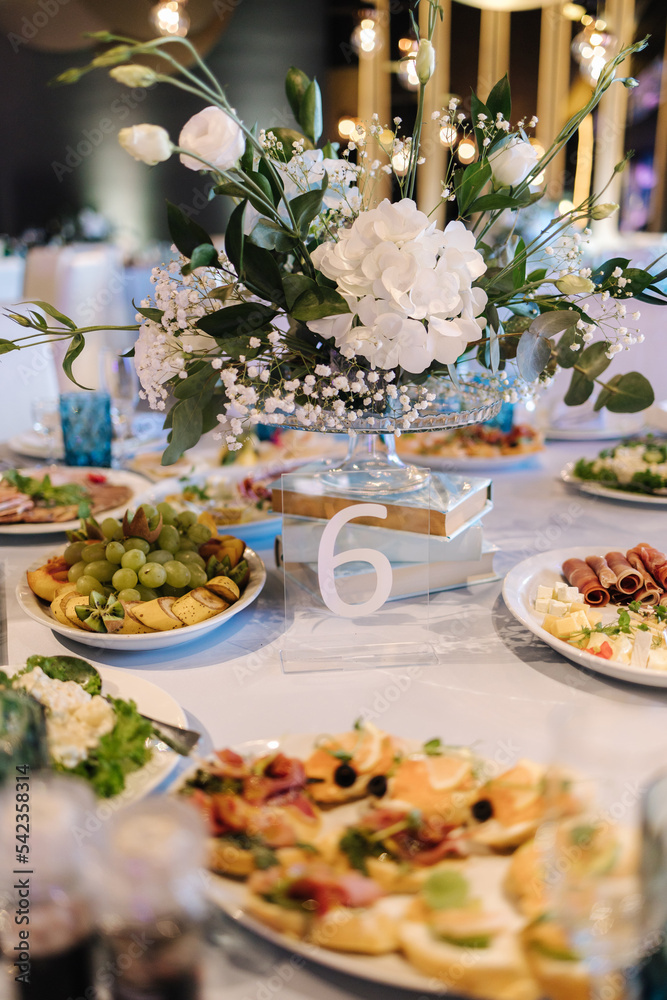 Big round table decorated by white and blue flowers in restaurant. Different dishes on table. Cristening of little boy. Luxury decor Big round table decorated by white and blue flowers in restaurant. Different dishes on table. Cristening of little boy. Luxury decor