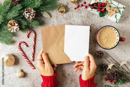 Desktop of woman hands in red sweater holding blank paper and envelope . Flat lay of gray background with cup of coffee and Christmas decoration. Top view mock up and copy space for text.