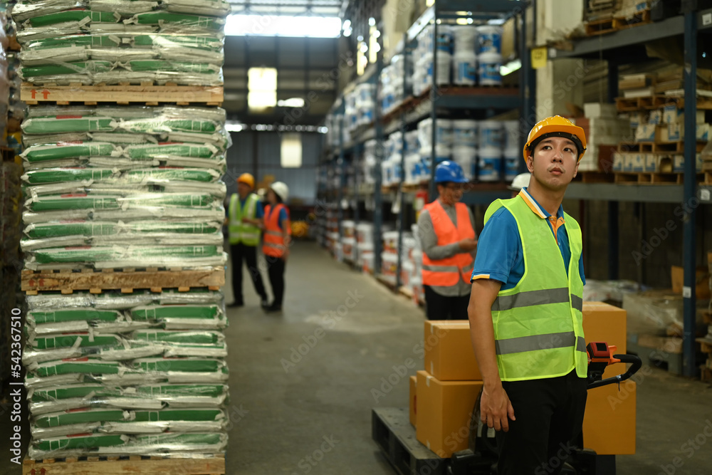 Shot of male warehouse worker in hardhats and jackets pulling a pallet truck walking through rows of storage racks with merchandise