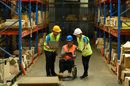Young worker and male manager in wheelchair using digital tablet, inspecting stock product in a large warehouse full of shelves