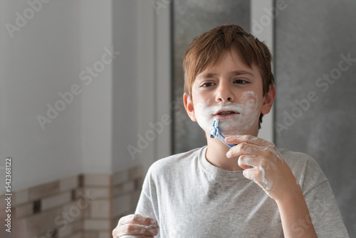 Boy imitates shaving beard with shaving foam on face like his father. Boy is fun in bathroom.